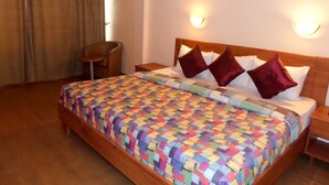 A hotel room with a wooden bed, a colorful quilt, two red pillows, a wooden nightstand, a chair, and an air conditioning unit on the wall. - Le Real Hotel (Lekki)