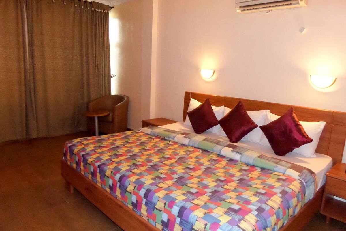 A hotel room with a wooden bed, a colorful quilt, two red pillows, a wooden nightstand, a chair, and an air conditioning unit on the wall.