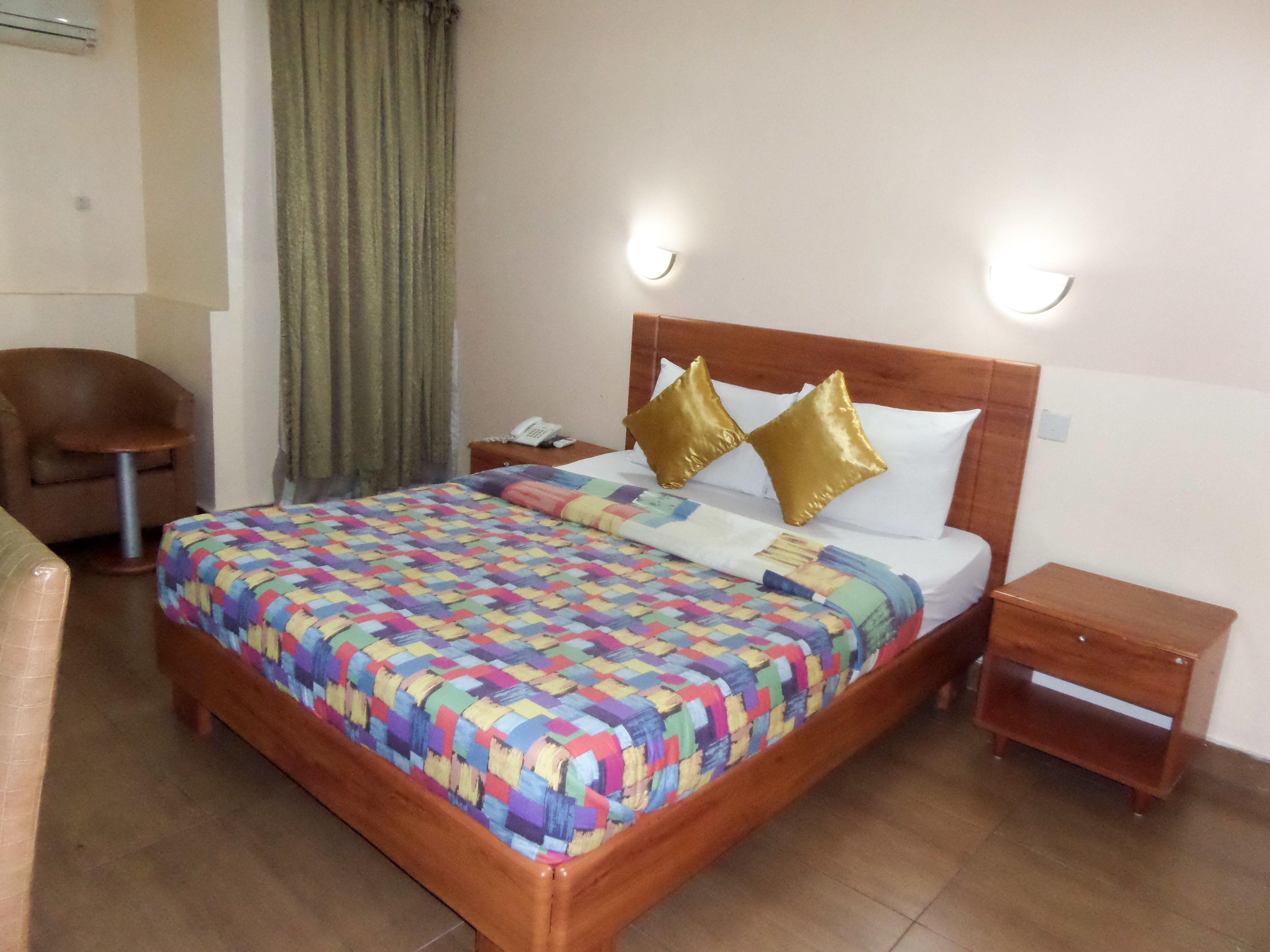 Een keurig opgemaakt bed met een kleurrijke quilt, twee gouden kussens en een houten hoofdbord. Er staan twee nachtkastjes, waarvan er één een telefoon heeft en de ander leeg is. Een bruine stoel is in de hoek te zien.