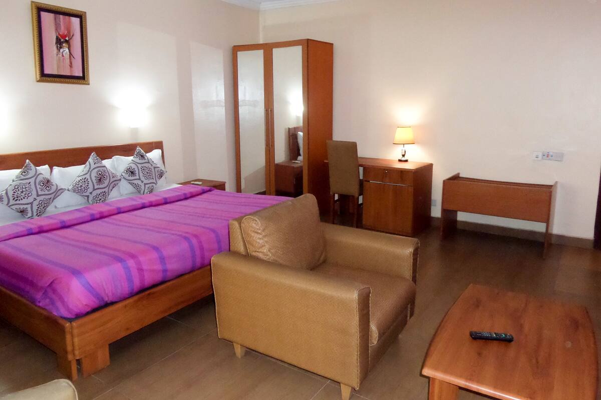 A hotel room with a bed, a chair, a desk, a lamp, and a remote control on a small table.