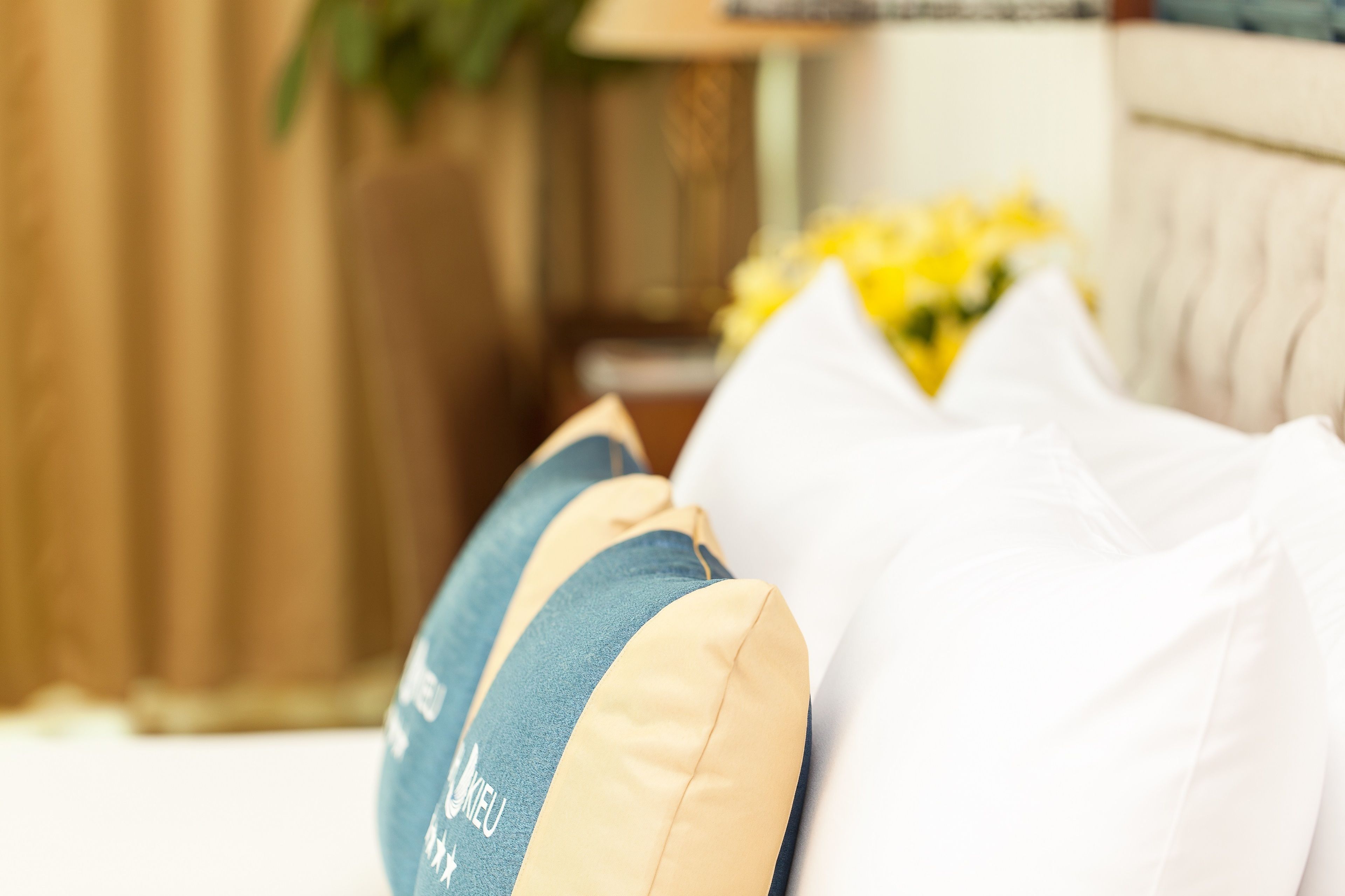 A close-up of a bed with multiple pillows, a white headboard, and a blurred background with a lamp and flowers.