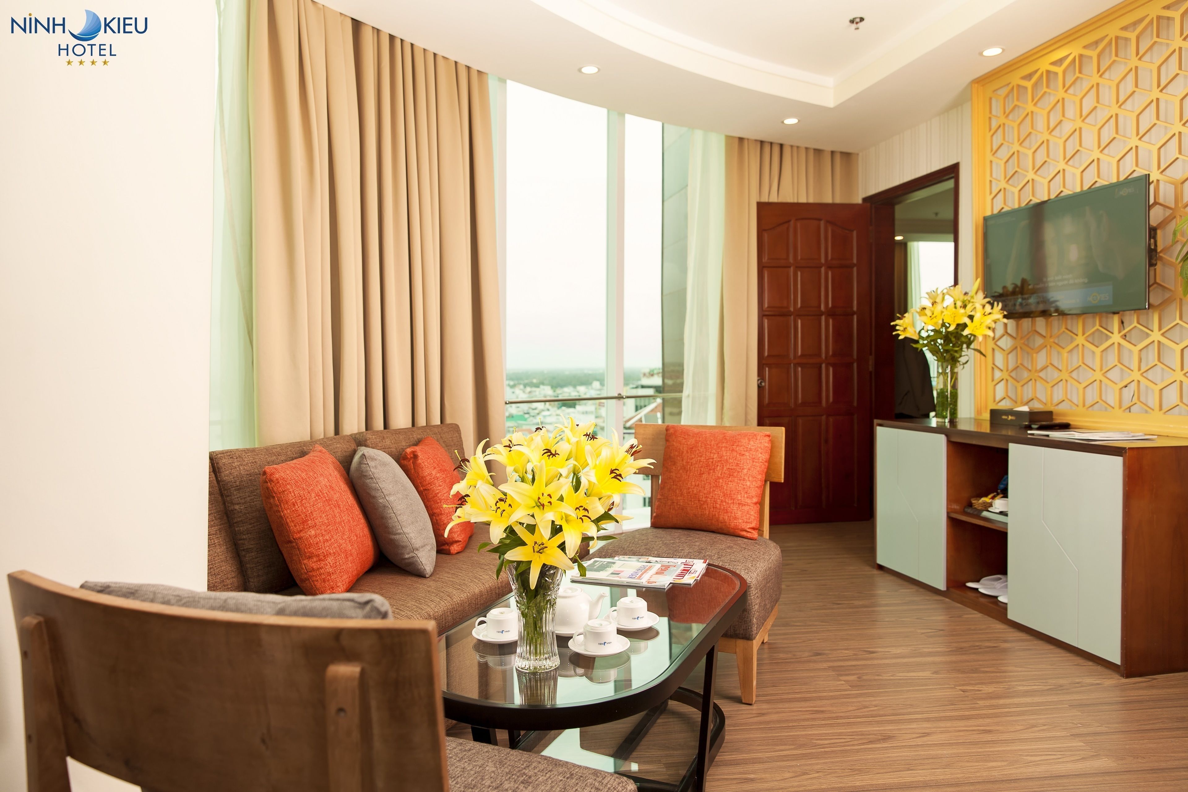 Habitación de hotel con sofá, una mesa de centro con un jarrón de flores, televisor y un mueble de madera.