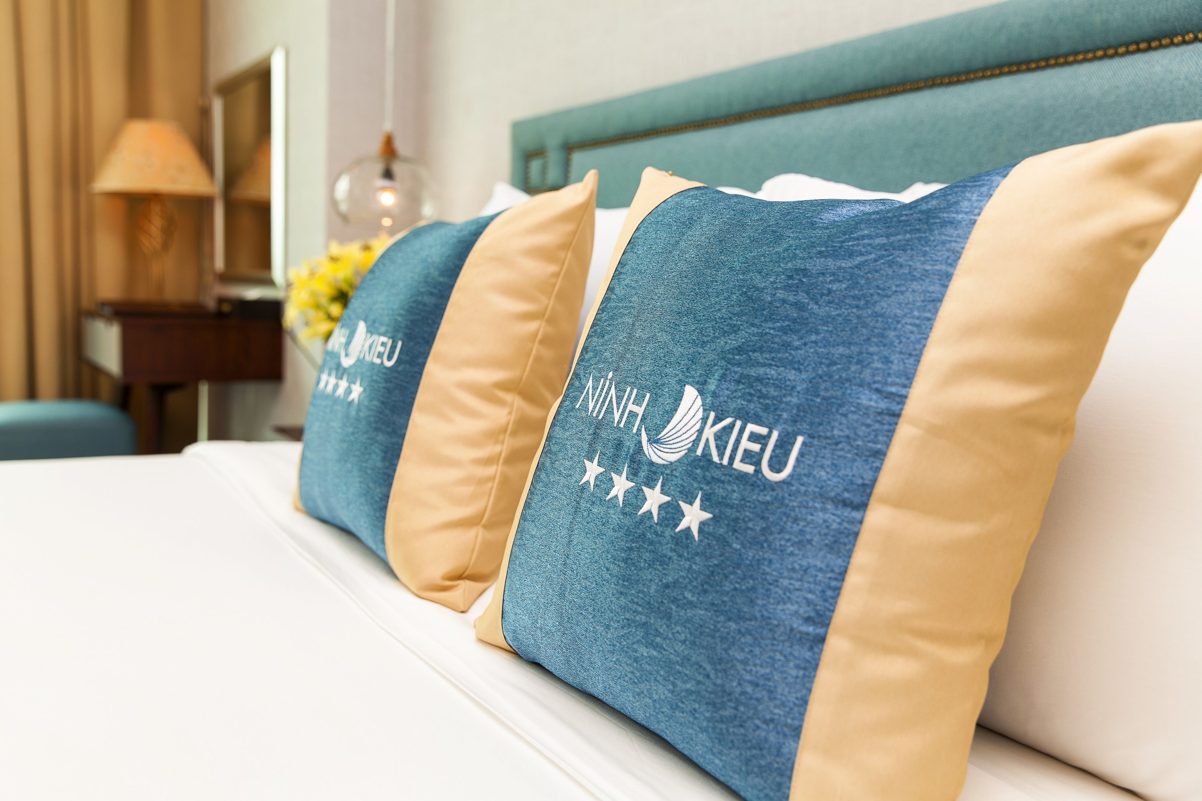 Un lit d’hôtel avec des oreillers bleus et beiges ornés du logo « MNH KIEU ».