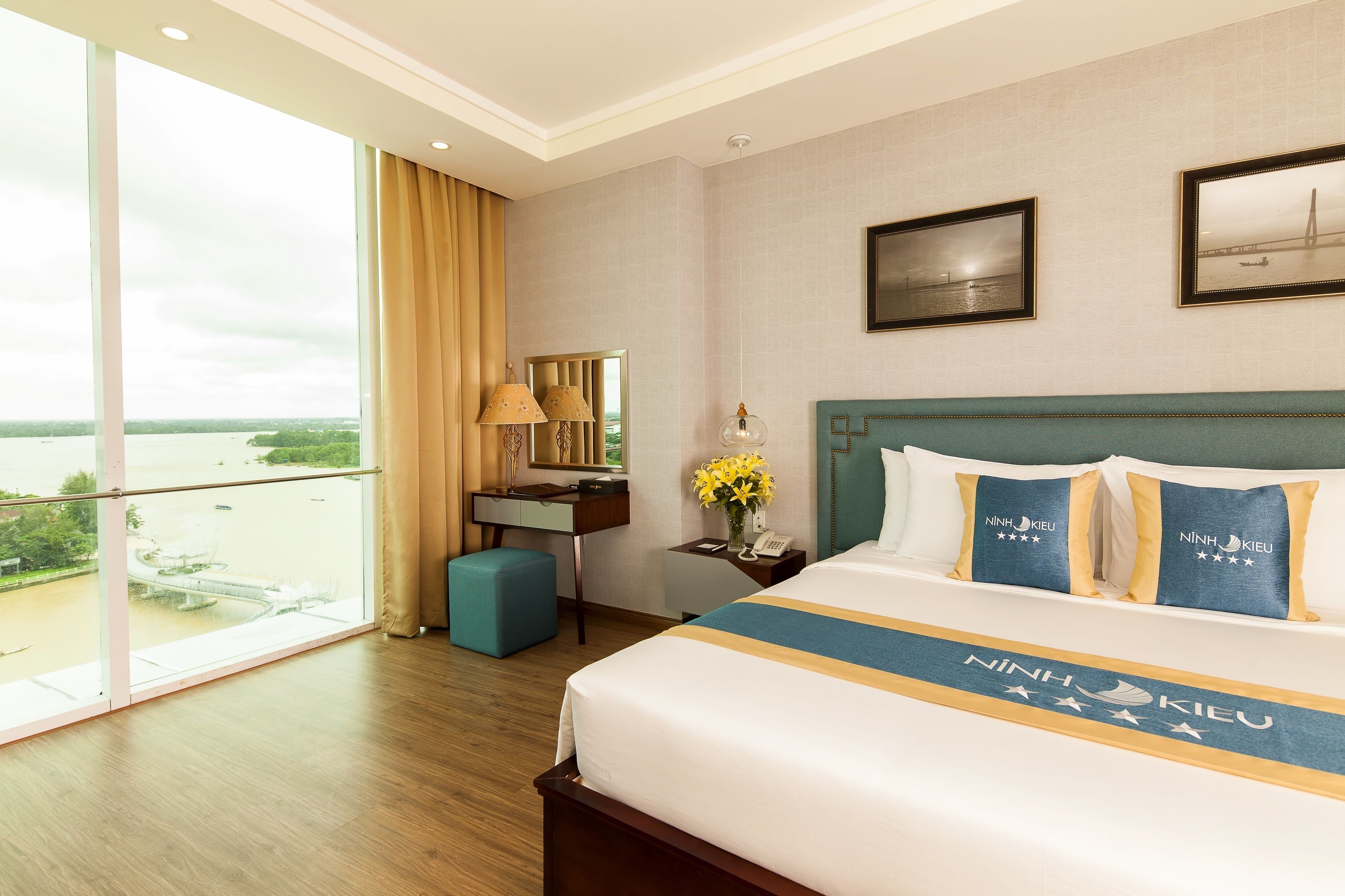 Habitación de hotel con un ventanal grande, una cama con almohadas de marca, un escritorio y una mesita de noche con un florero.