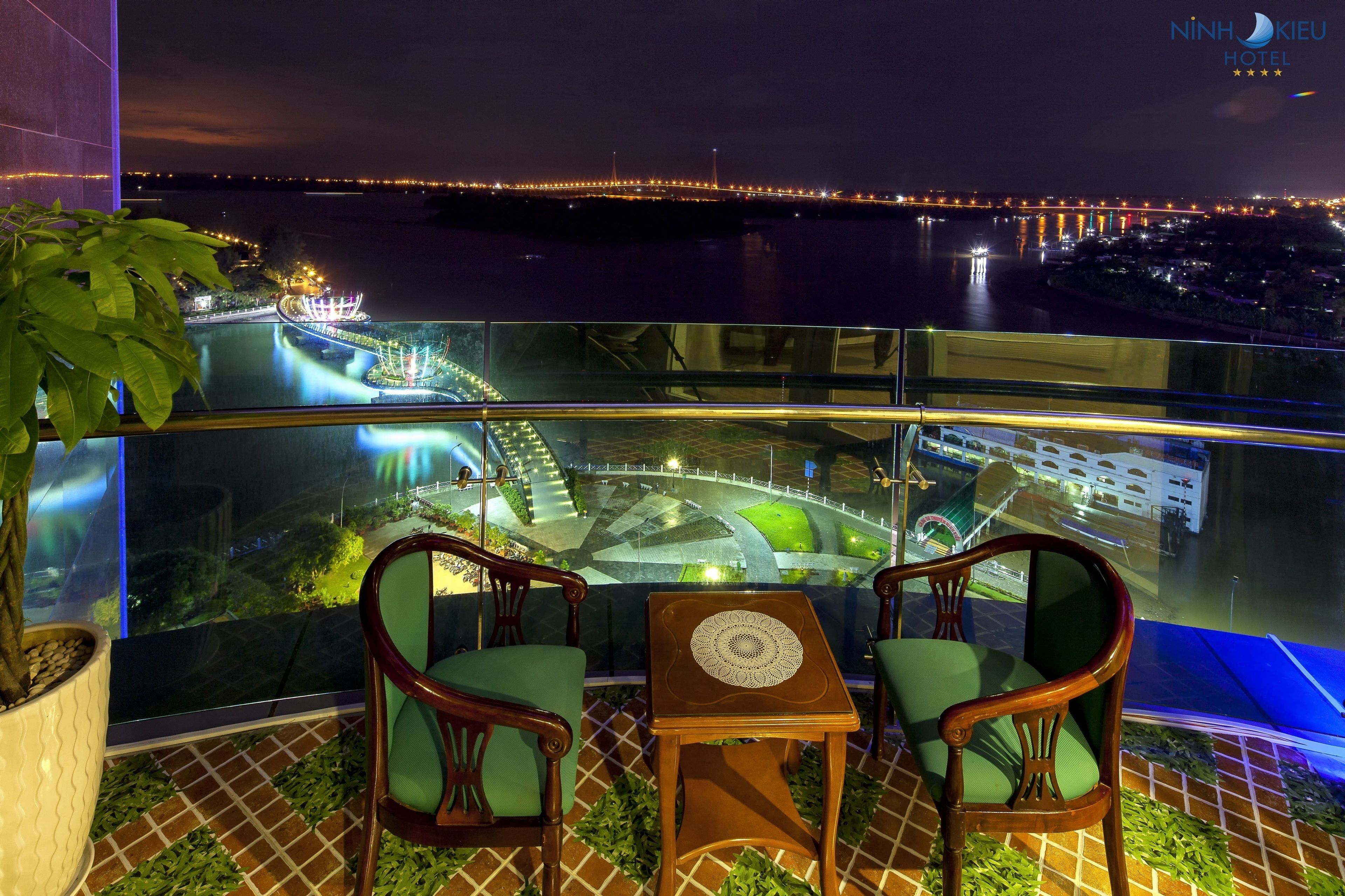 Un balcón con dos sillas de madera y una mesita, con vistas a un área ribereña bien iluminada que incluye un puente y edificios iluminados.
