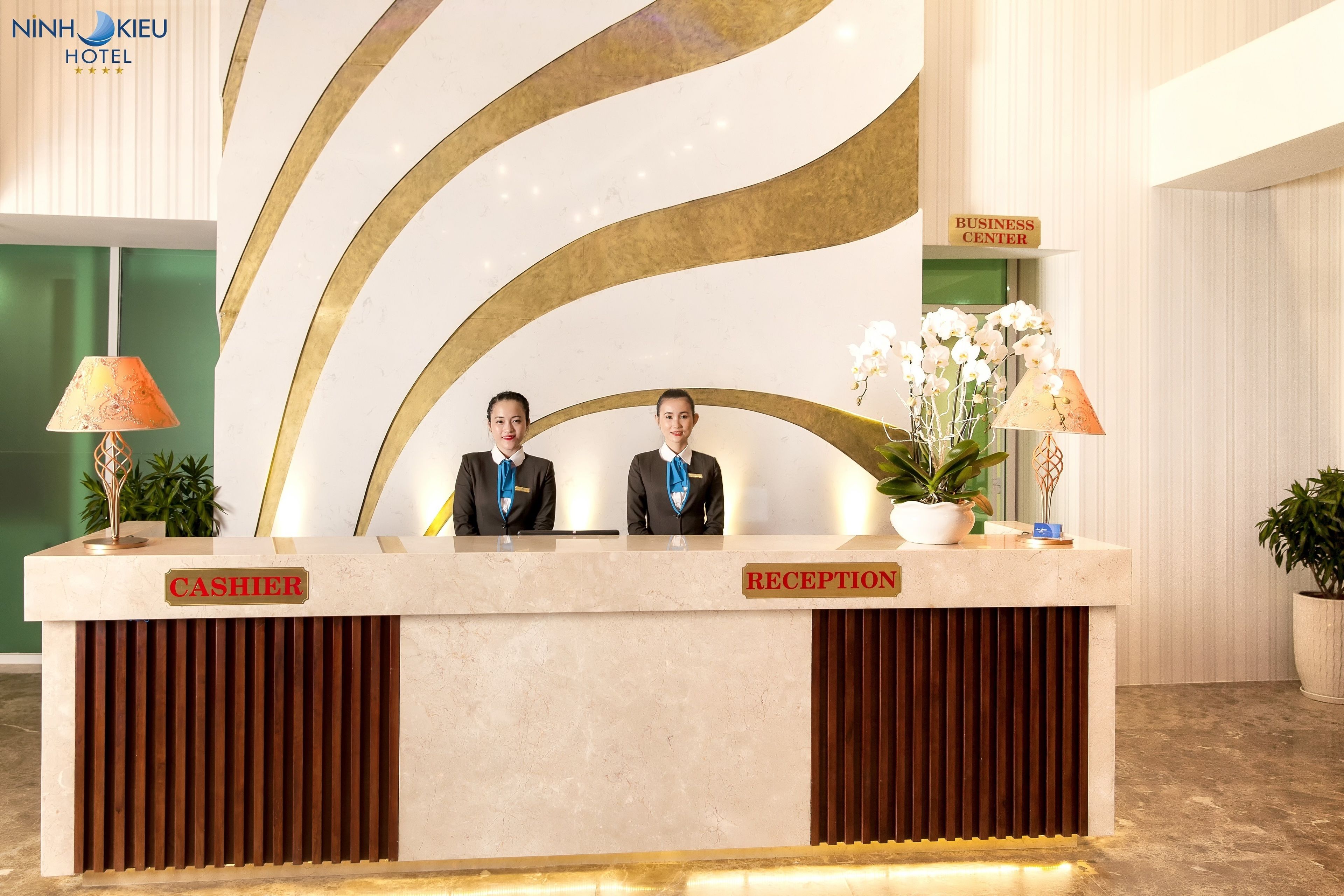 A hotel reception desk with a modern design, featuring a marble counter, two receptionists, and a sign indicating the business center.