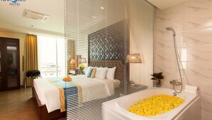 Una habitación de hotel moderna con una cama grande, un área de estar separada, una bañera llena de flores amarillas y una ducha.