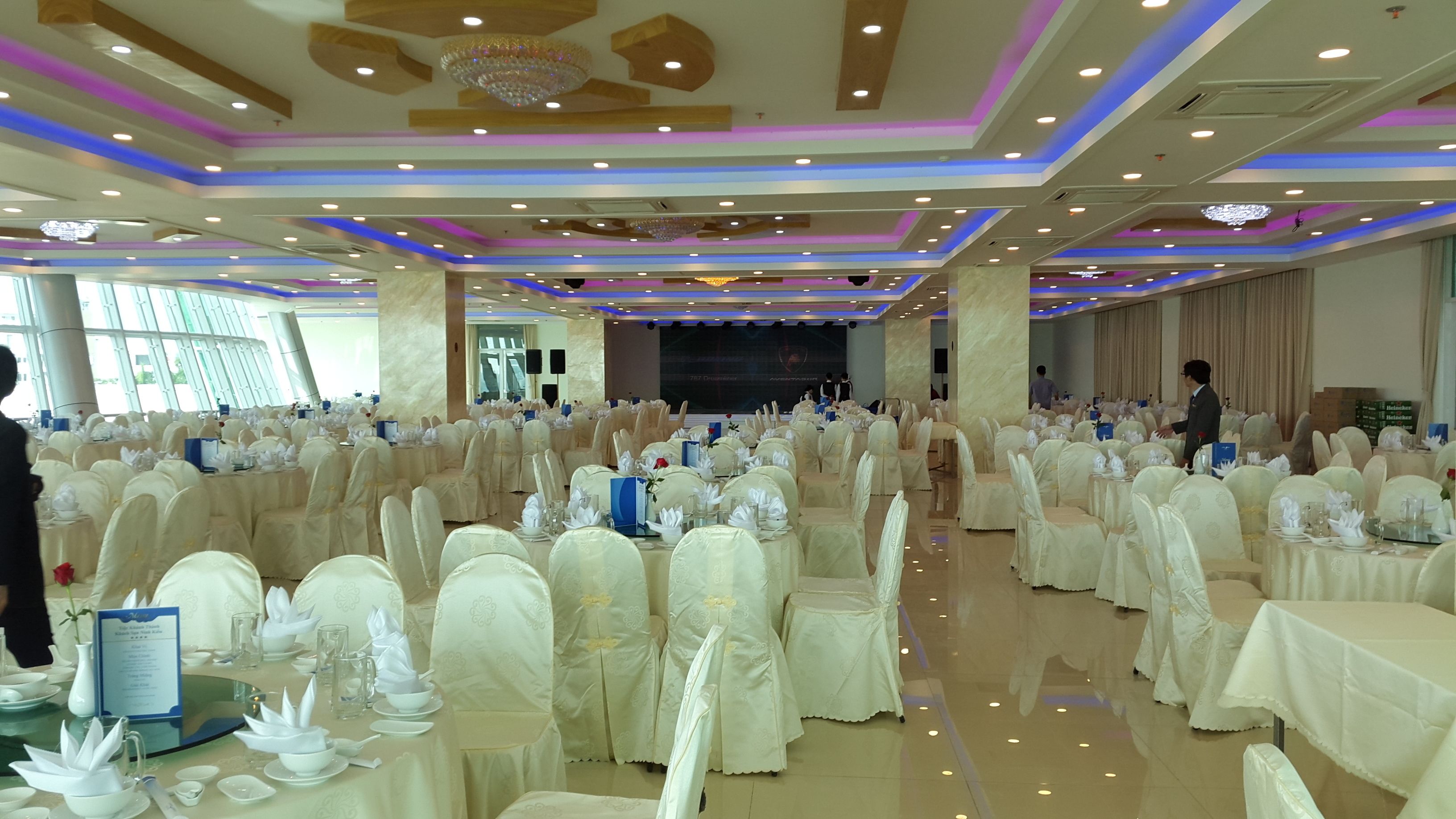 A banquet hall with round tables covered in white tablecloths and chairs, a stage with a large screen, and a chandelier.