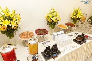 Una mesa de buffet con diversos alimentos, como frutas, pastelería y bebidas, y tres arreglos florales.