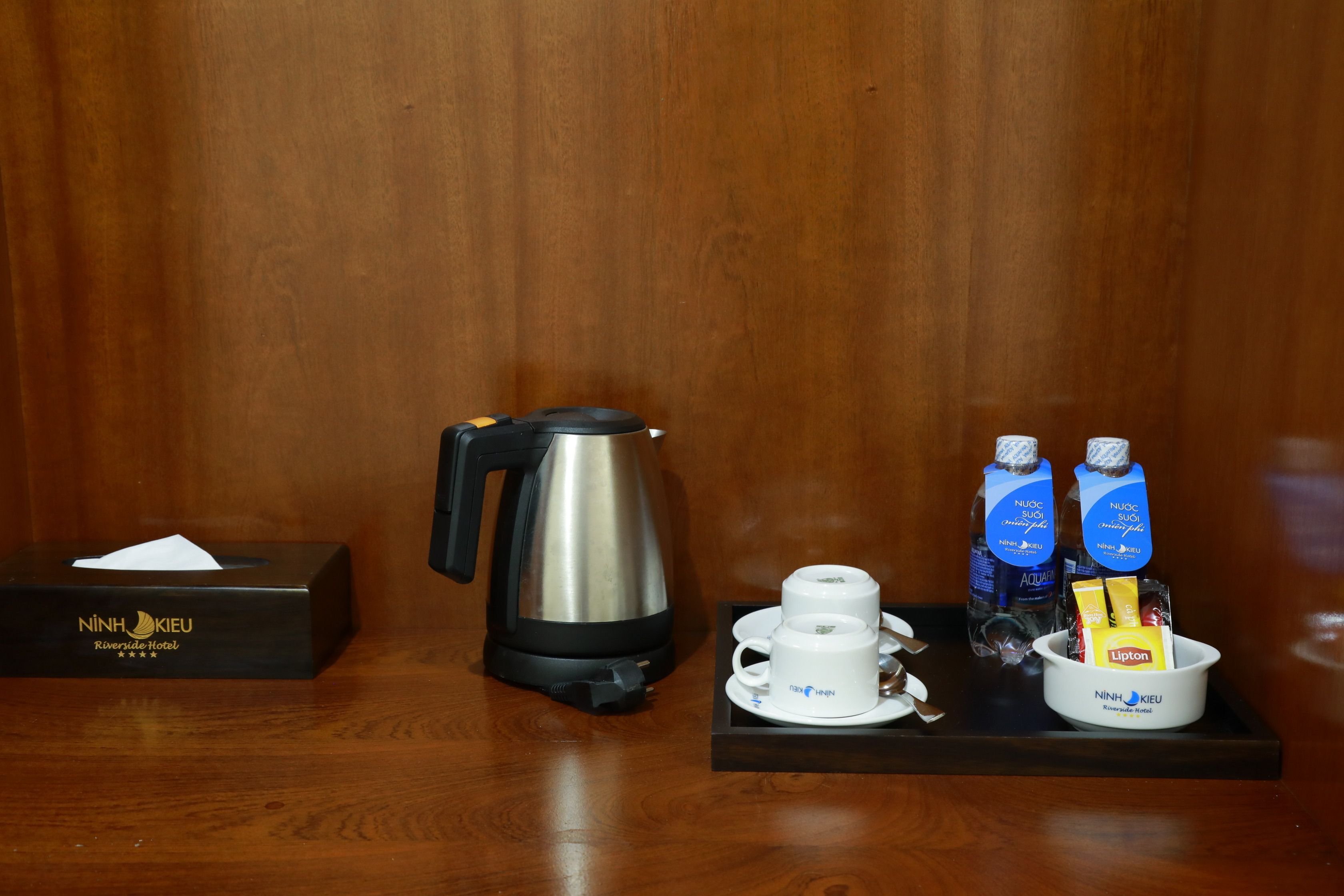 A hotel room with a wooden wall, a black tissue box labeled "NINH KIEU Riverside Hotel", a black electric kettle, a white cup and saucer, and a tray with two bottles of water, a Lipton tea bag, and a small bowl.