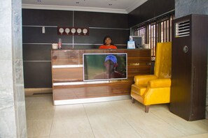 A reception area with a wooden reception desk, a television screen, a yellow armchair, and a clock on the wall. - Paris Suites (Lagos)