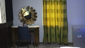 A hotel room with a bed, a desk with a chair, a mirror, a window with curtains, and an air conditioning unit.