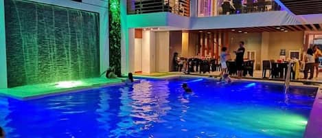 An indoor swimming pool with a visible logo, a seating area with tables and chairs, and a vertical garden feature.