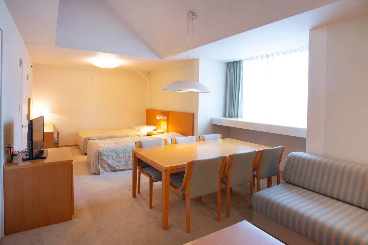 A hotel room with a bed, a desk with a computer, a dining table with chairs, a sofa, and a window with curtains.