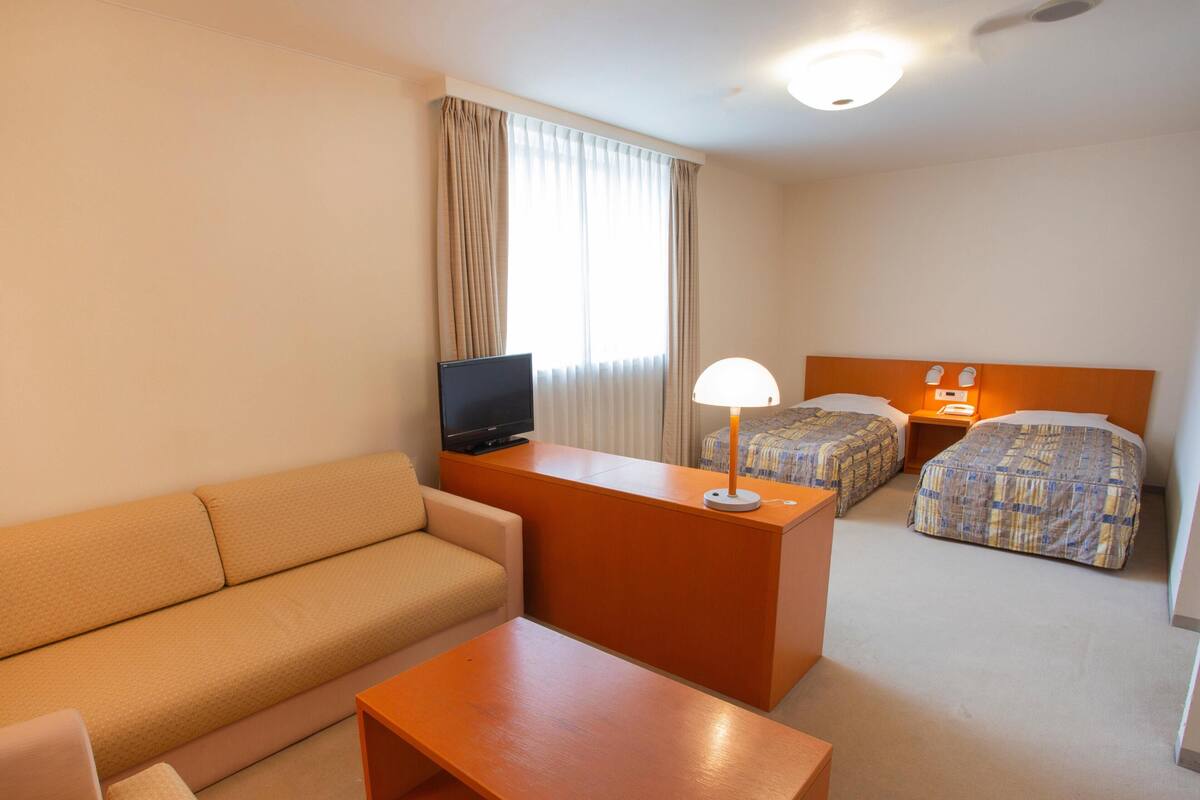 A hotel room with a sofa, a small table, a TV, two beds, and a nightstand with a lamp.