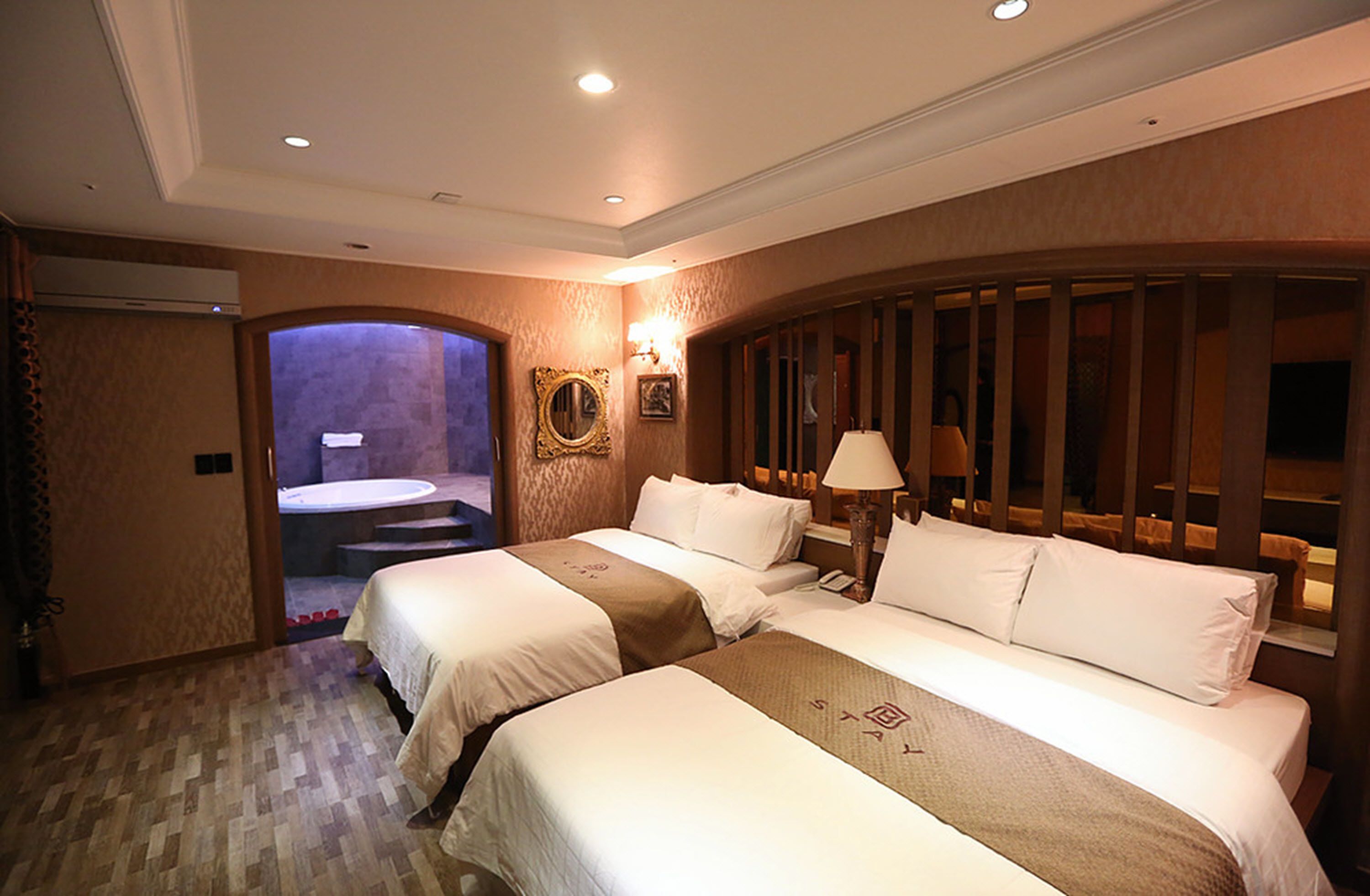 A hotel room with two beds, a bathroom with a shower, and a small sitting area with a lamp.
