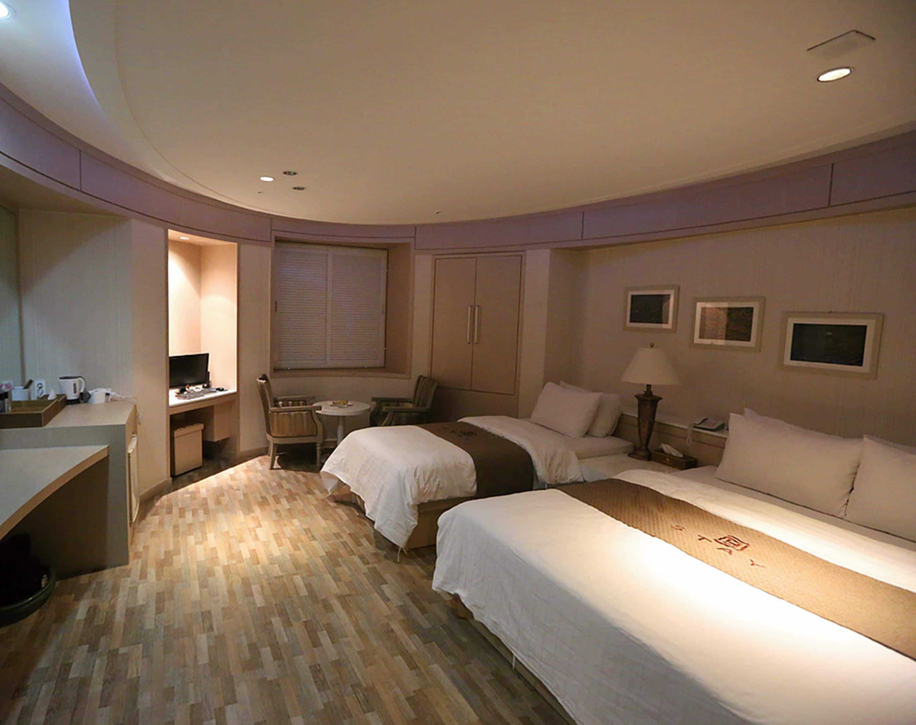 A hotel room with two beds, a desk, a chair, a small table, and a window with blinds.