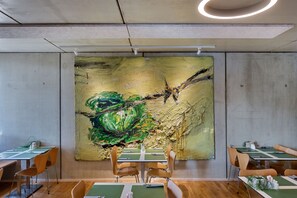 Un comedor con mesas y sillas, un cuadro abstracto grande en la pared y una lámpara en el techo.