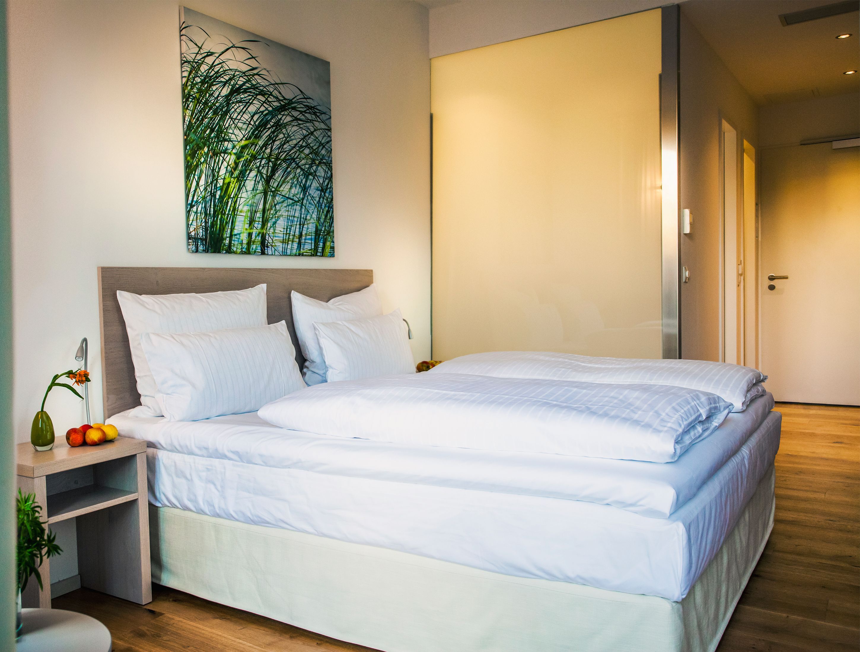 A neatly made bed with white linens, a bedside table with a vase of flowers, and a wall art piece above the bed.