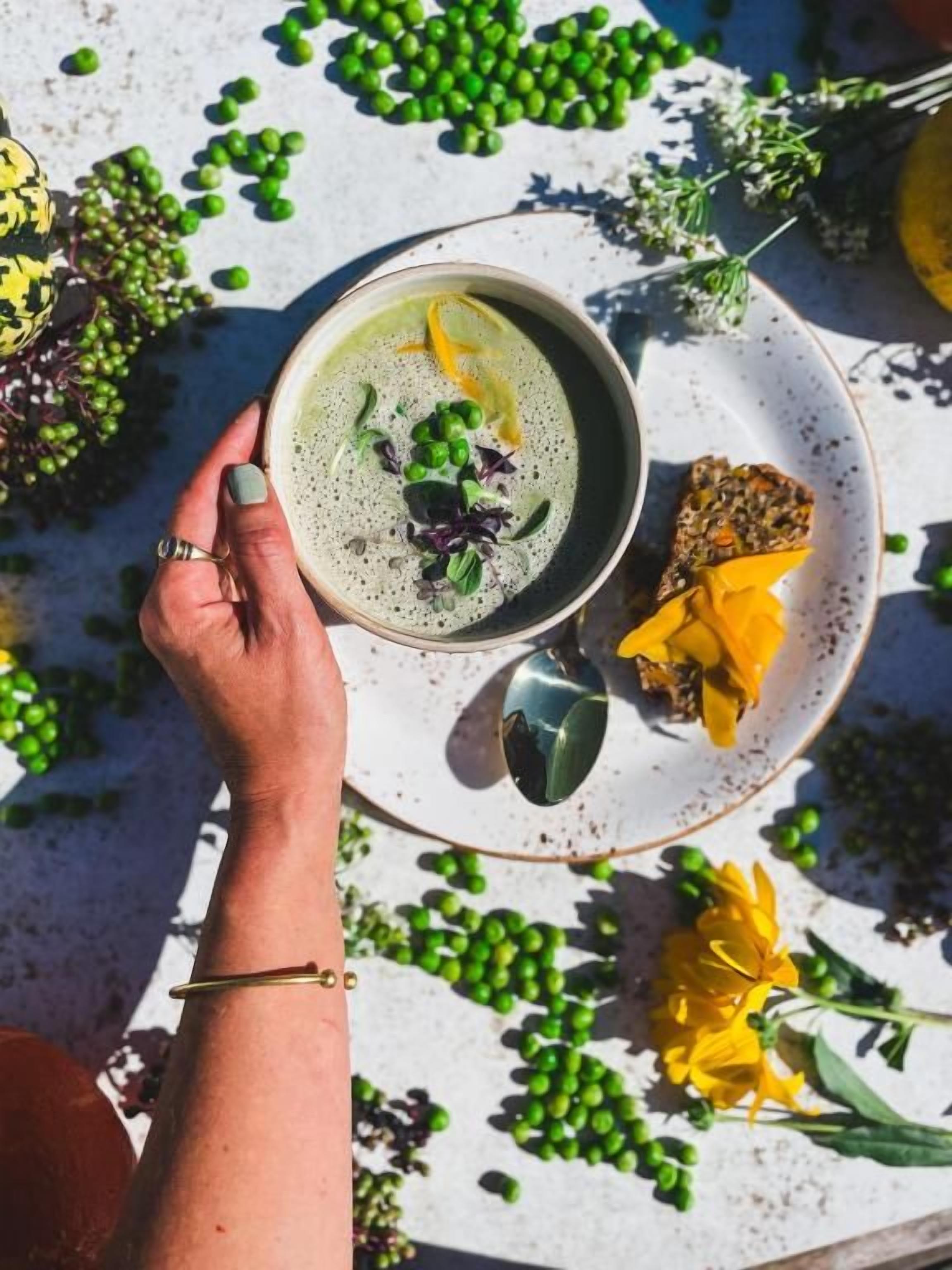 Uma pessoa segurando uma tigela de sopa, com um prato ao lado, cercada por ervilhas verdes e flores amarelas em uma mesa.