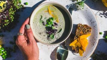 Uma pessoa segurando uma tigela de sopa, com um prato ao lado, cercada por ervilhas verdes e flores amarelas em uma mesa.