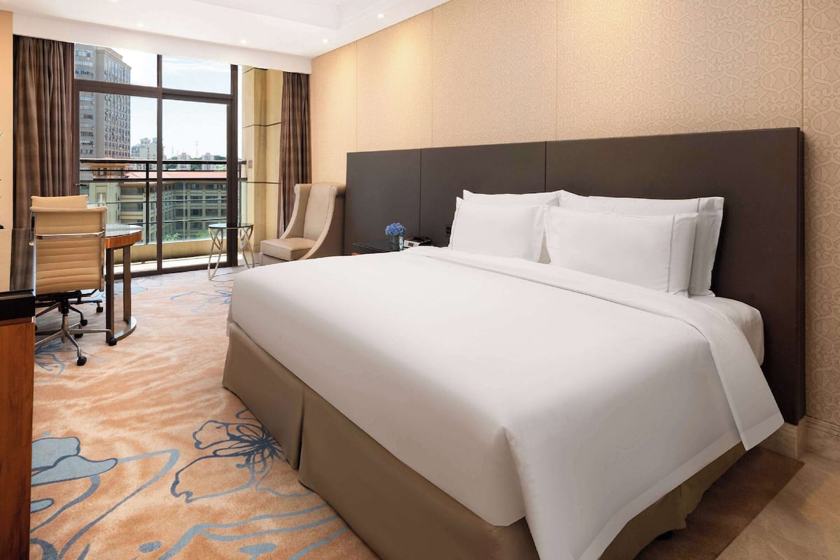 A hotel room with a large bed, a desk, a chair, and a view of the city through large windows.