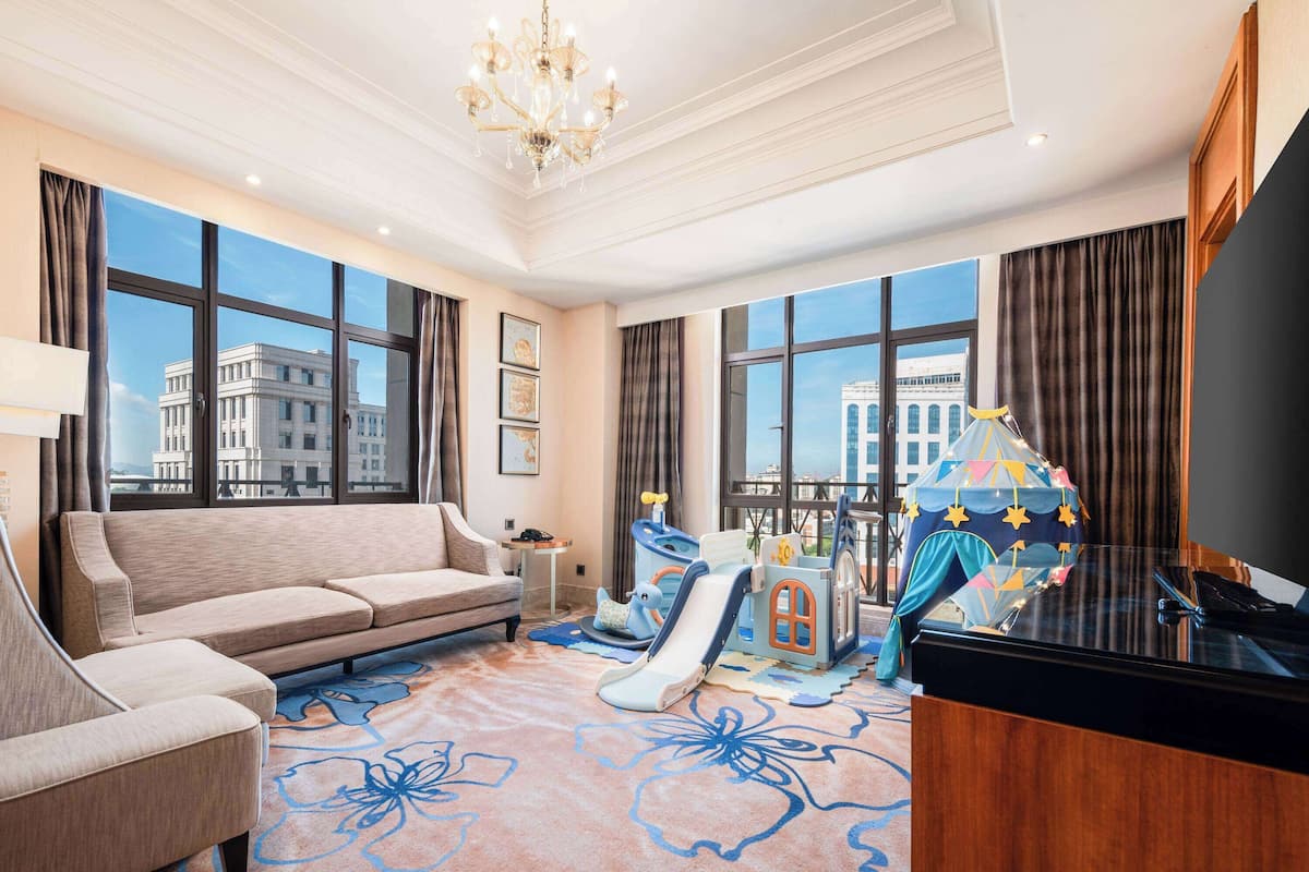 A spacious living room with a large sofa, a children's play area, a piano, and large windows offering city views.