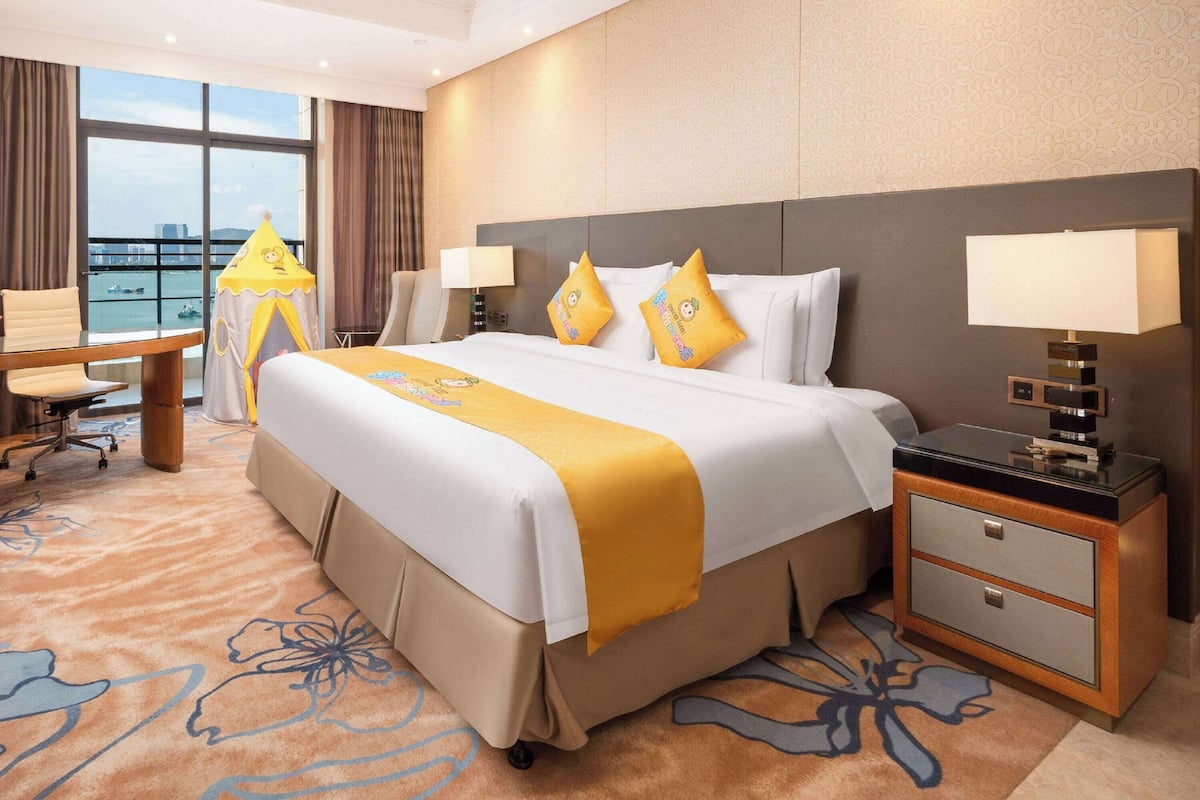 A hotel room with a large bed, a desk, a chair, a nightstand, and a view of the city through large windows.