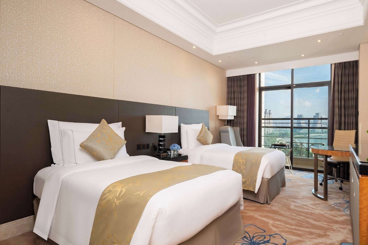 A hotel room with two beds, a desk, a chair, a lamp, and a view of the city.