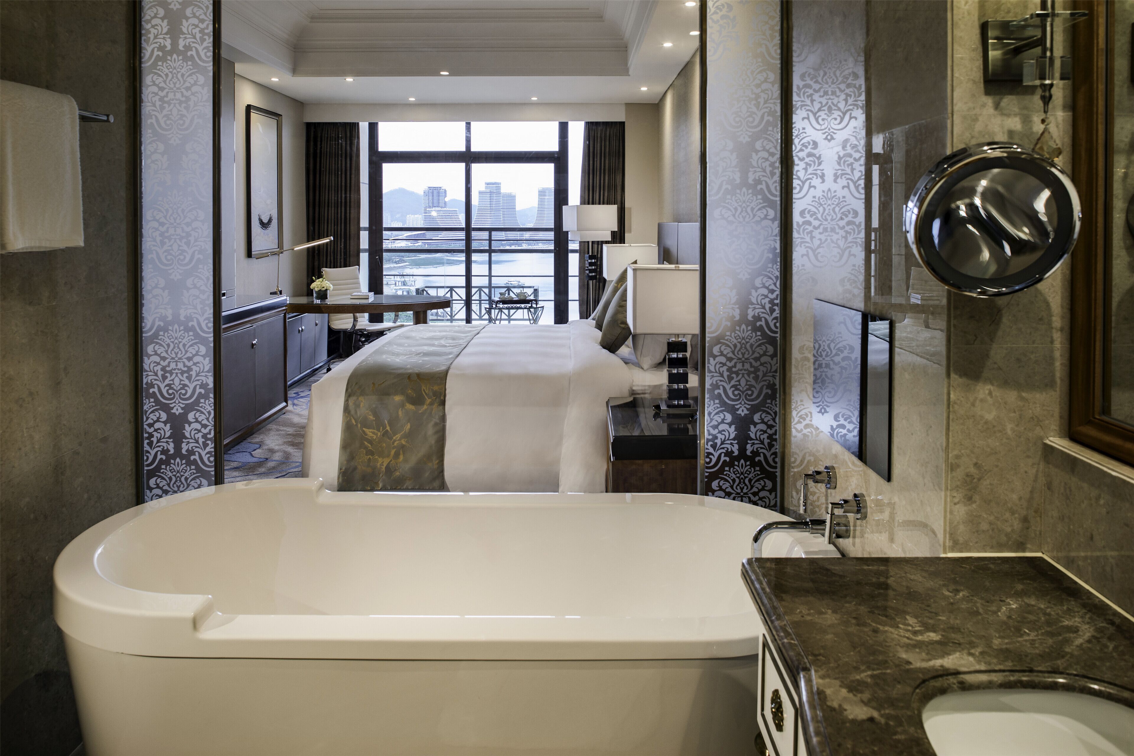 A modern hotel room with a large bathtub, a bed with a view of the city, a desk, and a bathroom with a large mirror.
