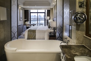 A modern hotel room with a large bathtub, a bed with a view of the city, a desk, and a bathroom with a large mirror.