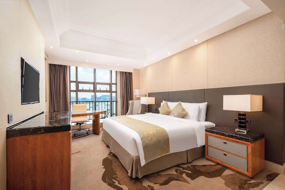 A hotel room with a large bed, a desk, a chair, a TV, and a view of the city.