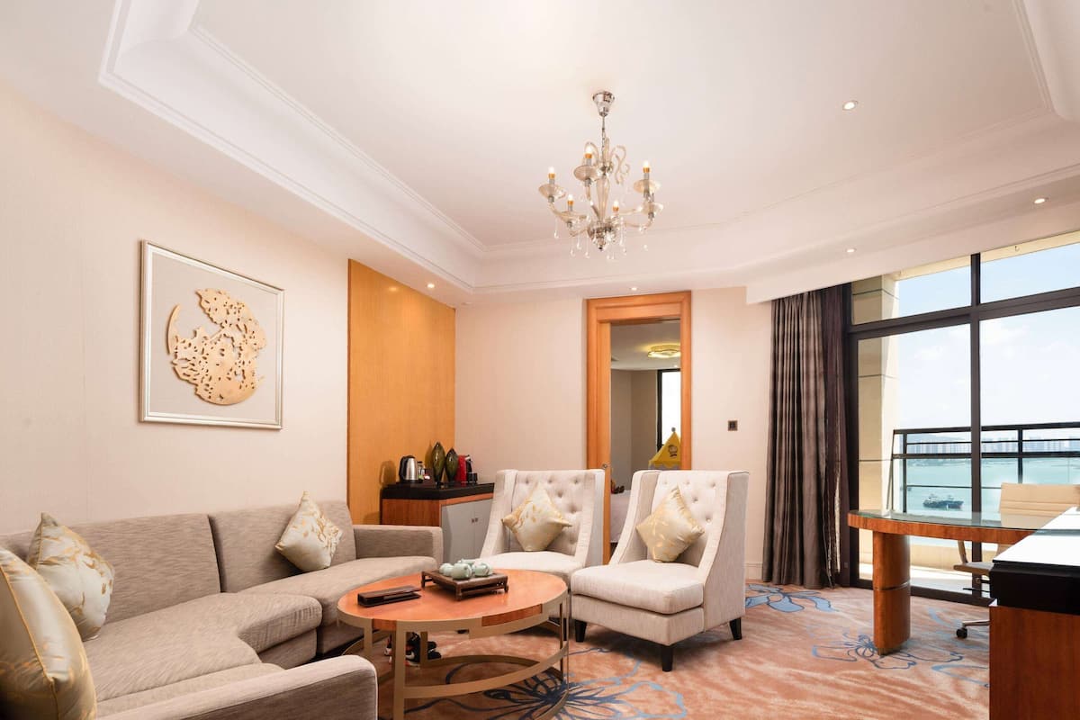 A hotel room with a sofa, two armchairs, a round coffee table, a chandelier, a large window with a view, and a framed artwork on the wall.