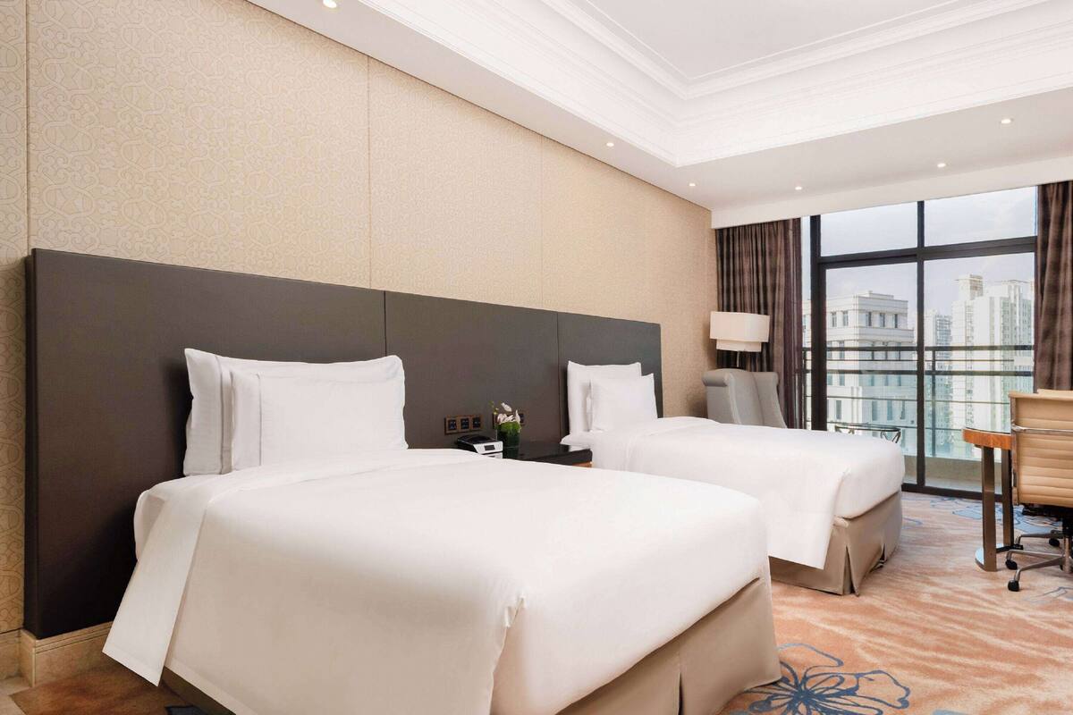 A hotel room with two beds, a large window with a city view, a desk, and a chair.