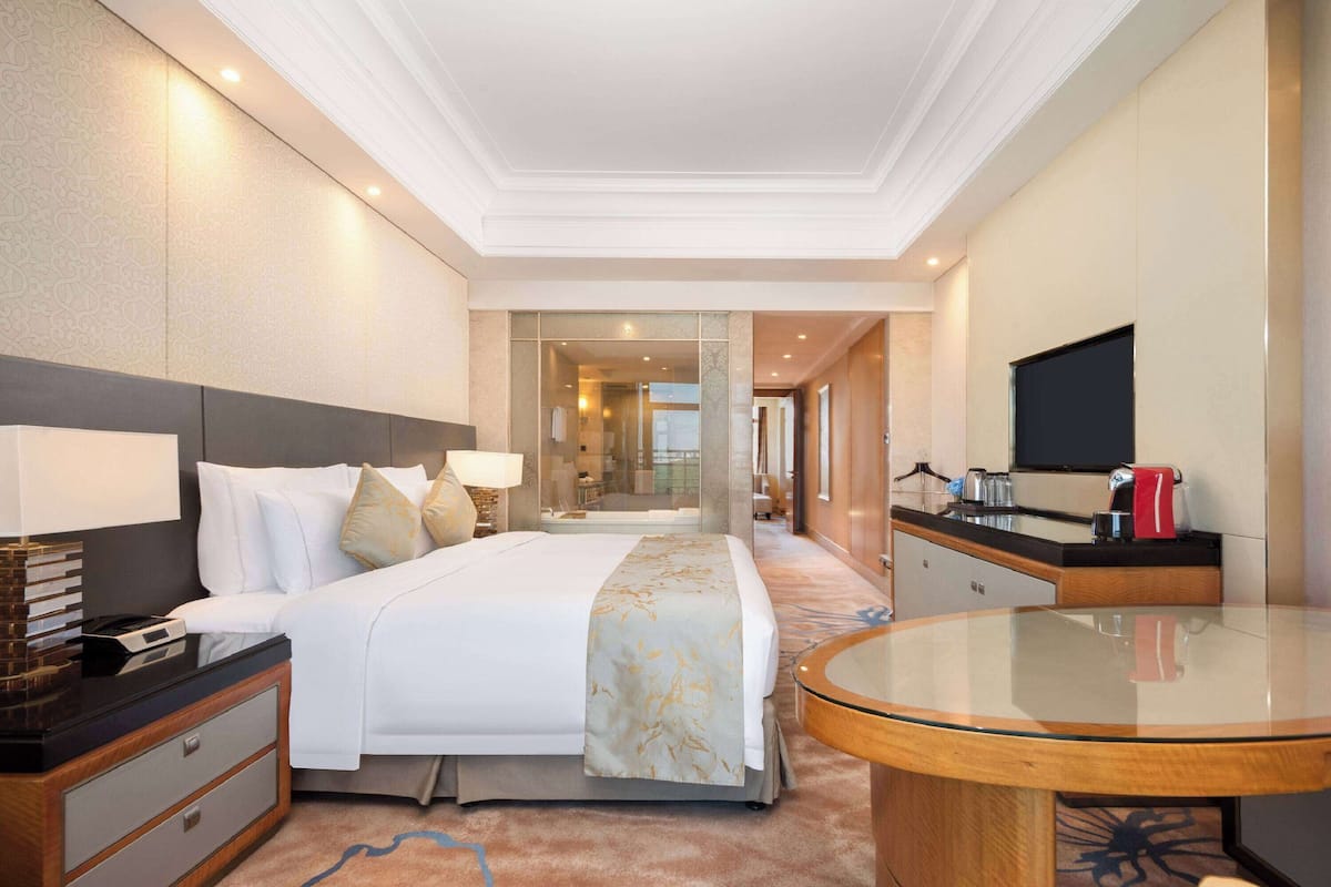 A hotel room with a large bed, a bedside table with a lamp, a round glass table, and a flat-screen TV mounted on the wall.