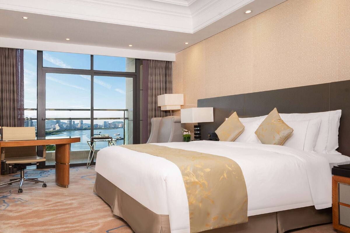 A hotel room with a large bed, a desk, and a view of a cityscape through large windows.