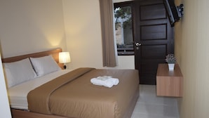 A neatly made bed with a brown bedspread and white pillows, a bedside lamp, a small table with a vase, and a dark wooden door with a window above it. - Roemah Canting (Yogyakarta)