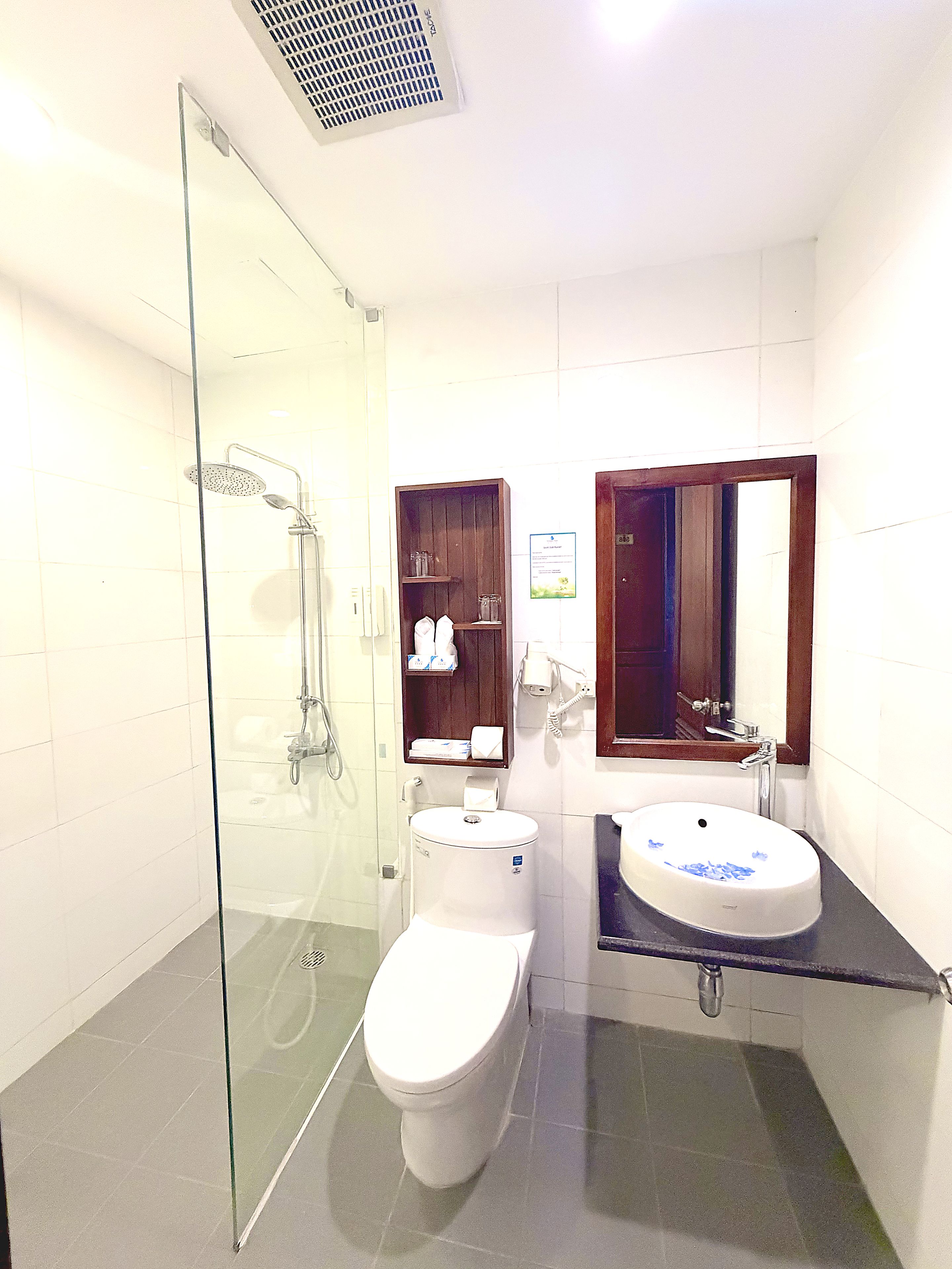 Un baño con ducha envuelta en vidrio, inodoro blanco, lavamanos con diseño floral azul y un mueble de madera sobre el lavamanos.