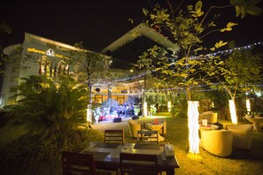Un área de comedor al aire libre con mesas y sillas, rodeada de vegetación e iluminación con luces colgantes.