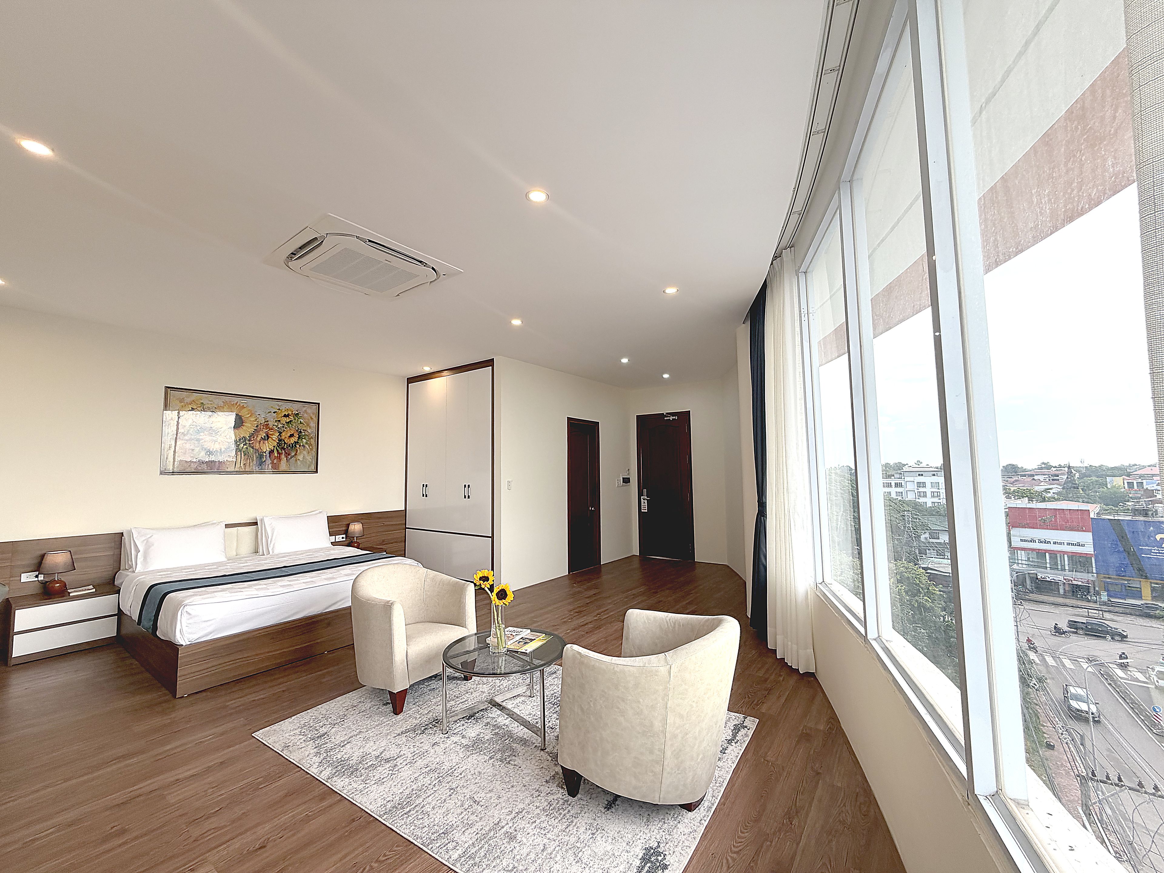 Una habitación de hotel moderna con una cama grande, una zona de estar con una mesa y sillas, un ventanal con vistas a la ciudad y una puerta que conduce a otra habitación.
