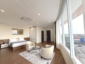 Une chambre d’hôtel moderne dotée d’un grand lit, d’un coin salon avec une table et des chaises, d’une grande fenêtre donnant sur la ville et d’une porte menant à une autre pièce.
