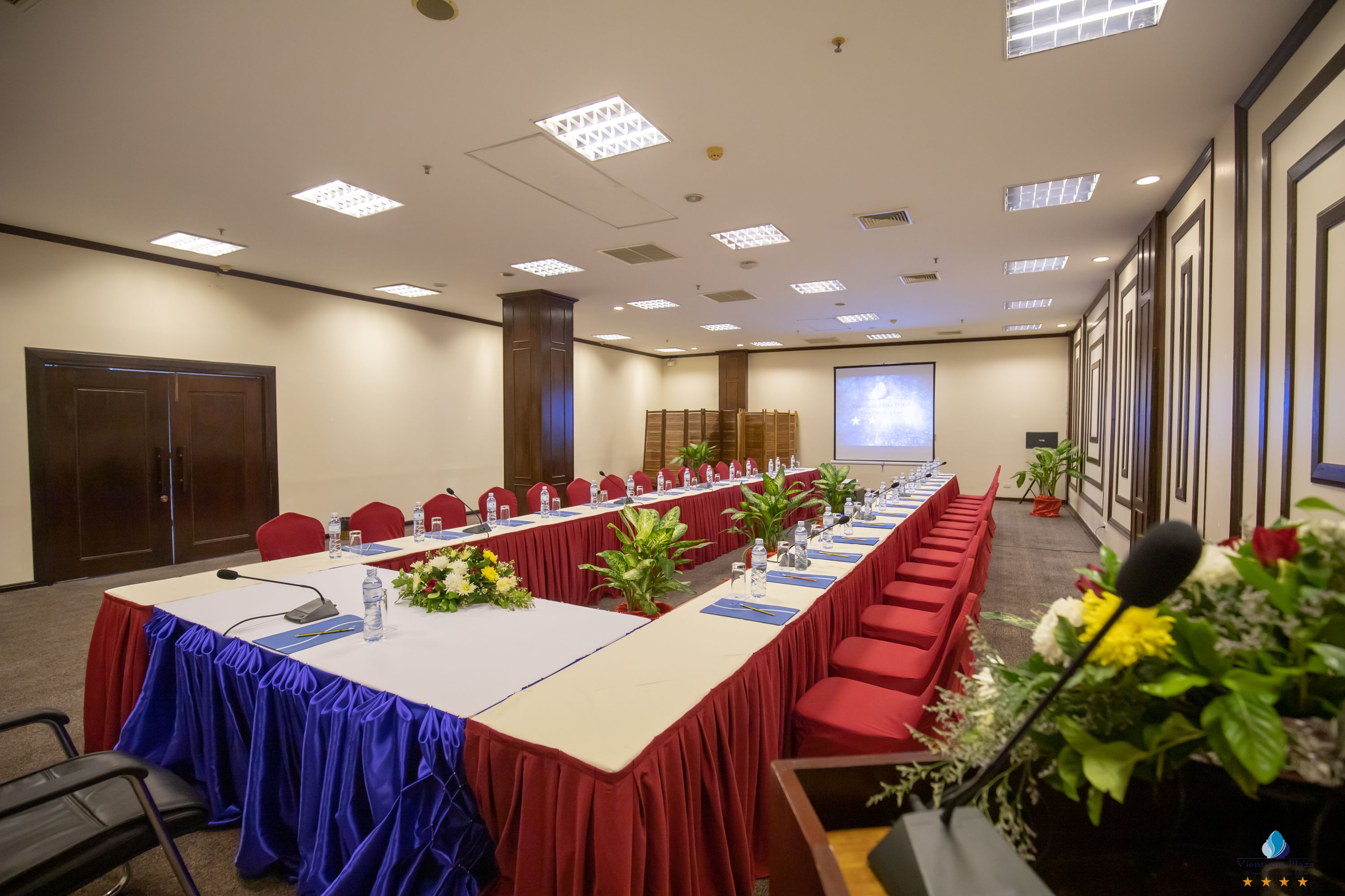 A conference room with a long table, red chairs, and a projector screen.