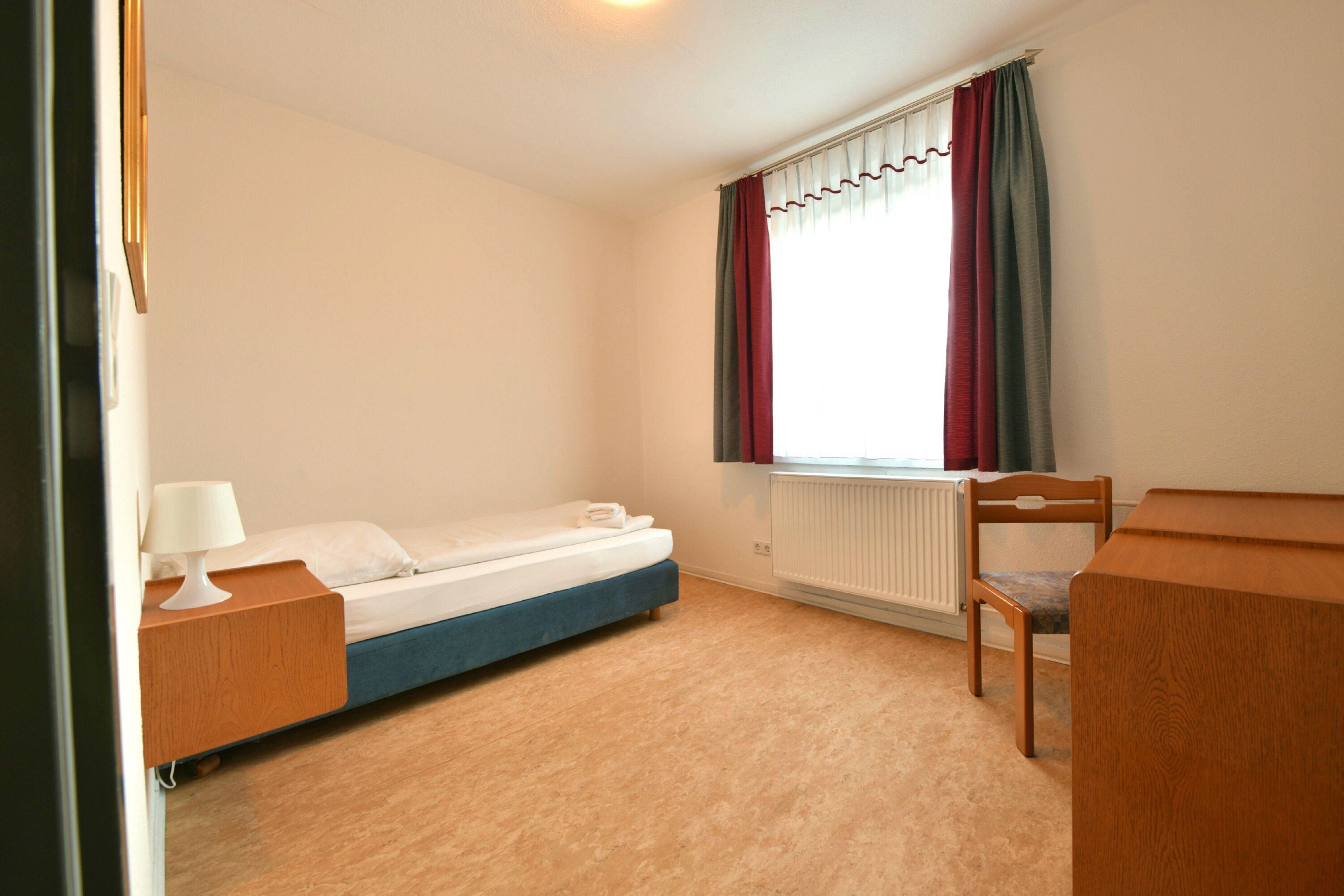 A small, clean room with a single bed, a wooden desk, and a chair.