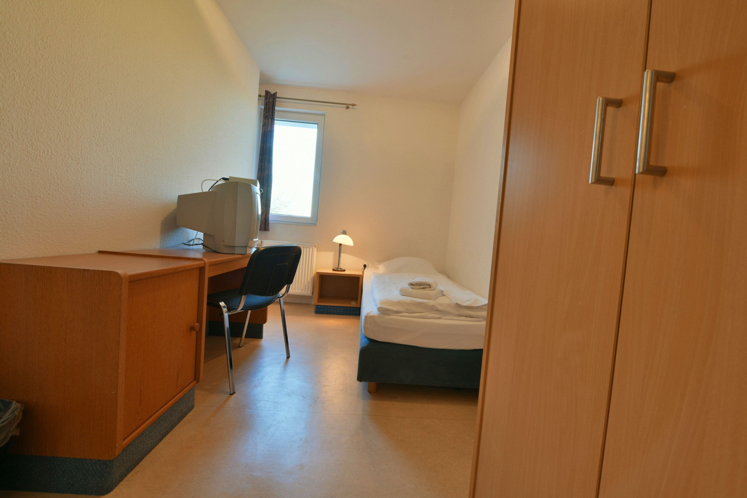 A compact hotel room with a wooden desk, a chair, a bed, and a nightstand with a lamp.