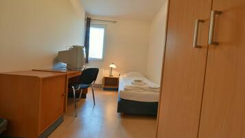 A compact hotel room with a wooden desk, a chair, a bed, and a nightstand with a lamp.