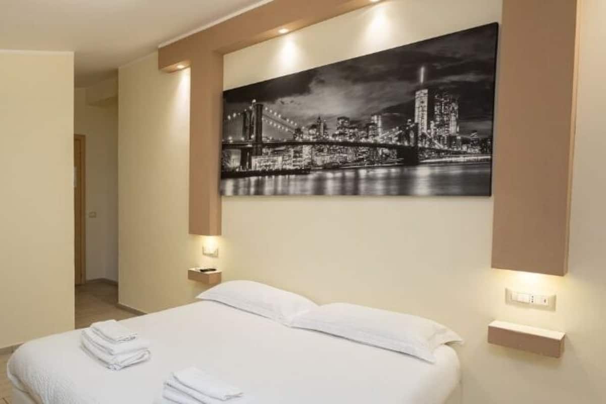 Ein Hotelzimmer mit einem Bett, einem Schreibtisch und einem Stuhl. An der Wand hängt ein Stadtbild.