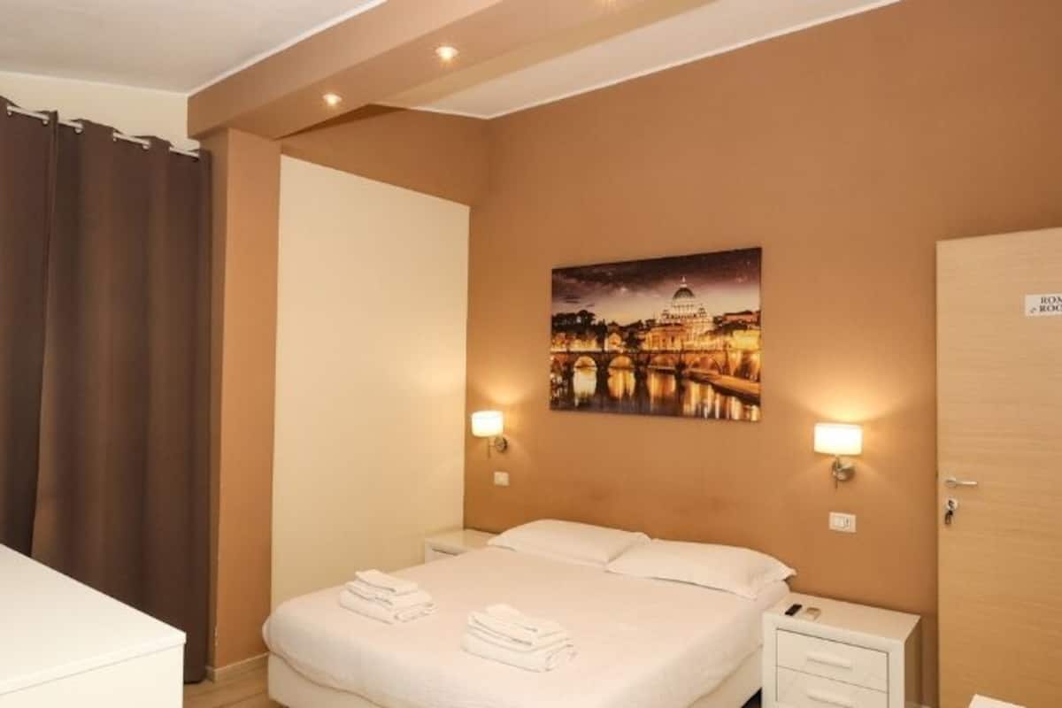 Ein Hotelzimmer mit einem Bett, einem Nachttisch, einer Lampe, einem Bild einer Brücke und einer Tür mit der Aufschrift 'Rome Room'.