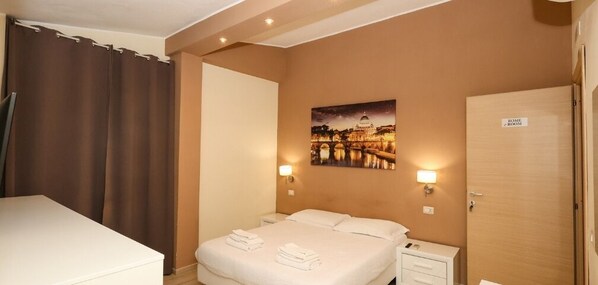 Quarto de hotel com uma cama, mesa de cabeceira, luminária, pintura de uma ponte e uma porta com a inscrição 'Rome Room'.