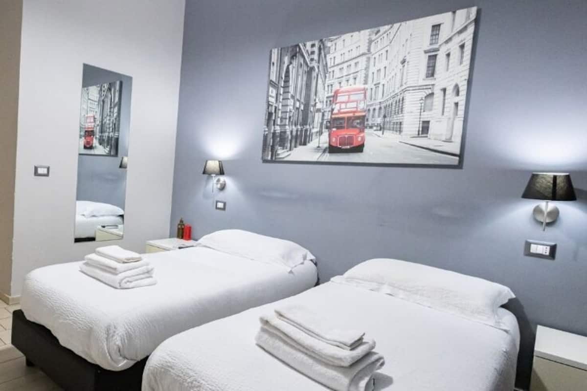 Ein Hotelzimmer mit zwei Betten, einem Schreibtisch und einem Stuhl. An den Wänden sind Lampen angebracht und ein gerahmtes Bild eines roten Doppeldeckerbusses hängt dort.