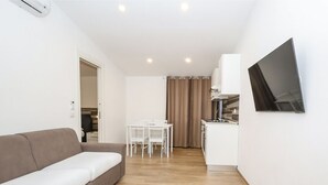 Un espace de vie compact comprenant un canapé, une table à manger avec des chaises, un coin cuisine avec une cuisinière et un téléviseur à écran plat fixé au mur.
